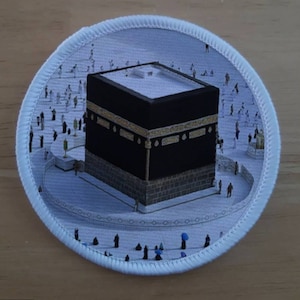 Puede incluir: Un parche circular blanco con una imagen negra y dorada de la Kaaba, un edificio en forma de cubo en La Meca, Arabia Saudita. La Kaaba está rodeada de un área blanca con muchas figuras pequeñas de personas.