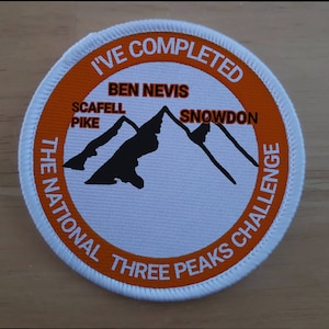 Puede incluir: Un parche redondo blanco y naranja con el texto "I've Completed The National Three Peaks Challenge" y una silueta negra de tres montañas etiquetadas como "Ben Nevis", "Scafell Pike" y "Snowdon".