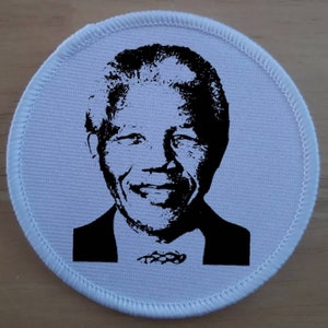 May include: A white round fabric patch featuring a black and white image of Nelson Mandela. The image shows Mandela's face and shoulders, with a smile on his face. The patch has a white border with a stitched edge.