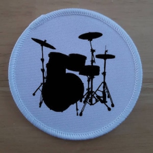 May include: A white round patch with a black silhouette of a drum set. The drum set includes a bass drum, two tom-toms, a snare drum, and two cymbals.