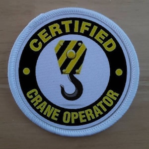 May include: White round patch with black and yellow border. The center of the patch has a white background with a black crane hook and the text "CERTIFIED CRANE OPERATOR".