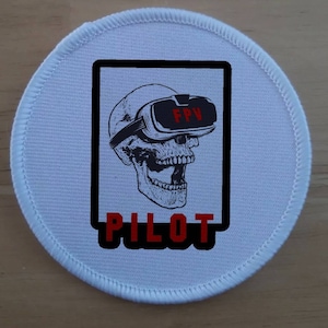 Könnte beinhalten: Runder weißer Patch mit schwarzem Rand und einem Schädel, der ein Virtual-Reality-Headset trägt, mit dem Text "FPV PILOT" in Rot.