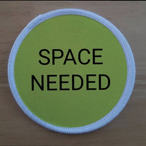 May include: A round, green fabric patch with white stitching around the edge. The text "SPACE NEEDED" is printed in black on the patch.