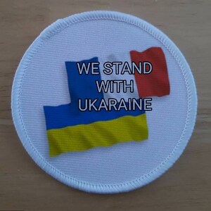 May include: A white round patch with a blue and yellow Ukrainian flag and a red flag. The text "WE STAND WITH UKRAINE" is printed on the patch.