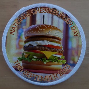 Pode incluir: Um remendo circular branco com uma borda vermelha e amarela. O remendo apresenta um grande cheeseburger com alface, tomate, queijo e um picles. O texto "National Cheeseburger Day 18th September 2023" é impresso ao redor da borda do remendo.
