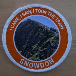 Może przedstawiać: Okrągła naszywka w kolorze białym i pomarańczowym z tekstem "I came, I saw, I took the train Snowdon" wokół zdjęcia góry.