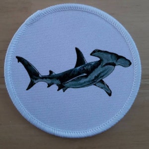 May include: Circular patch with a detailed illustration of a hammerhead shark in grey and black. The shark is set against a light blue background, with a white stitched border. Suitable for application on jackets, bags, or other fabric items.