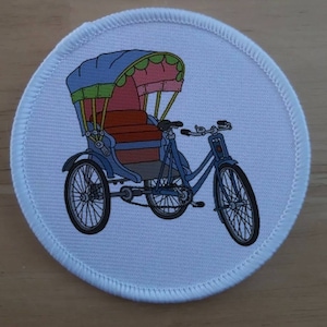 May include: A white fabric patch with a colorful illustration of a traditional Asian rickshaw. The rickshaw has a blue canopy with pink and green trim, a brown seat, and three wheels.