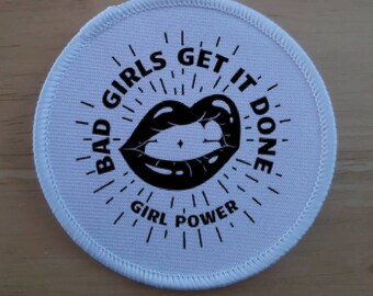 May include: A circular, white patch featuring a black lip graphic and the words "BAD GIRLS GET IT DONE" and "GIRL POWER" in a radiating design. The patch has a stitched border.