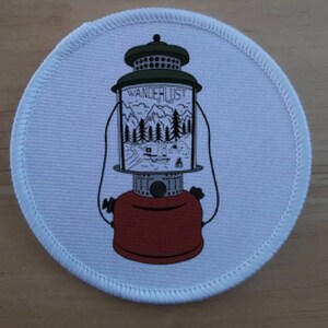 May include: White round patch with a black and white illustration of a vintage lantern with the word "Wanderlust" printed on the side. The lantern has a red base and a green top.