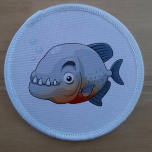 May include: Round patch featuring a cartoon piranha with a gray body, blue fins, and a red belly. The piranha has a wide grin with sharp teeth and a friendly expression. The patch has a white border.