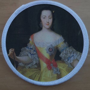 Könnte beinhalten: Ein runder gestickter Aufnäher mit dem Porträt einer Frau in einem gelben und weißen Kleid mit einer roten Schärpe. Sie trägt eine Perlenkette und Ohrringe. Der Hintergrund ist dunkel.