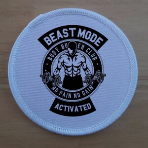 Peut inclure: Patch rond blanc avec du texte noir et une silhouette noire d'une personne musclée soulevant des poids. Le texte indique "Beast Mode Body Builder Club No Pain No Gain Activated".