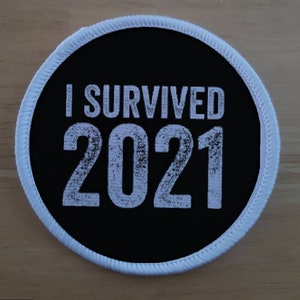 Può includere: Un cerotto rotondo nero e bianco con cuciture bianche attorno al bordo. Il cerotto dice "I Survived 2021" in testo bianco.
