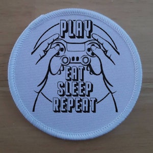 May include: White round patch with black text and a graphic of a hand holding a video game controller. The text reads "Play Eat Sleep Repeat."