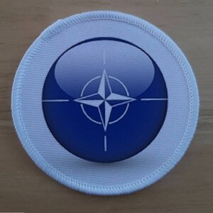 May include: A white round patch with a blue and white NATO symbol in the center. The symbol is a compass with a star in the center.