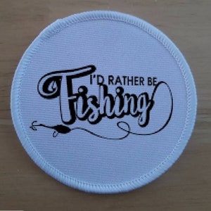 Peut inclure: Écusson rond en tissu blanc avec du texte noir qui dit "I'd rather be fishing" avec un hameçon et une ligne de pêche stylisés.