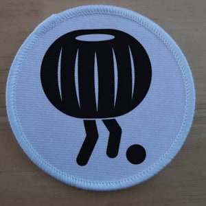 May include: White round fabric patch with a black cartoon image of a round object with legs and a small black circle.