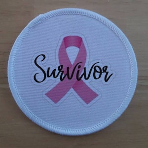 May include: White round fabric patch with a pink ribbon and the word "Survivor" in black cursive writing.