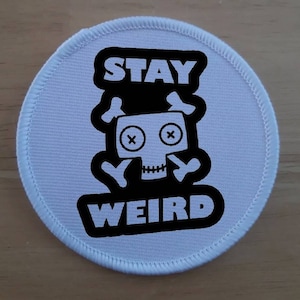 Puede incluir: Parche redondo blanco con texto negro que dice "STAY WEIRD" y un gráfico de calavera en blanco y negro.