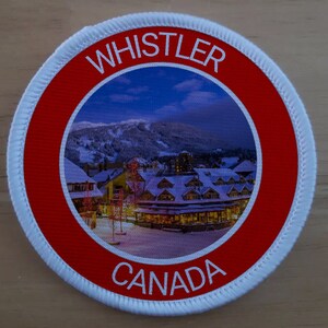 May include: A white round patch with a red border. The centre of the patch features a photo of a snowy mountain village at night. The text "WHISTLER" is printed in white on the red border above the photo. The text "CANADA" is printed in white on the red border below the photo.