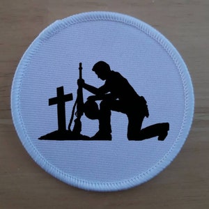 May include: A white round patch with a black silhouette of a soldier kneeling at a grave with a rifle and a cross.