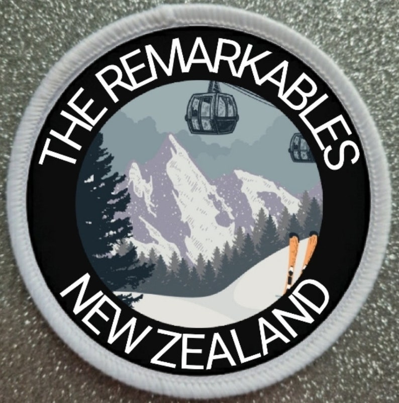 May include: A white circular patch with a black border and the text "THE REMARKABLES NEW ZEALAND" in white. The center of the patch features a stylized image of a snowy mountain range with a ski lift and skiers.