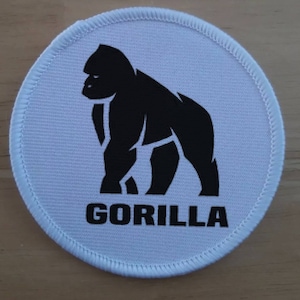 Könnte beinhalten: Ein runder, weißer Stoffaufnäher mit einer schwarzen Silhouette eines Gorillas im Profil. Unter der Tiergrafik ist das Wort GORILLA in fetten, schwarzen Großbuchstaben aufgedruckt. Der Aufnäher hat einen weiß umkettelten Rand.