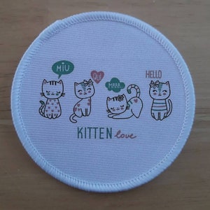 Könnte beinhalten: Ein runder Stoff-Patch in Weiß mit vier Comic-Katzen und dem Text "KITTEN love". Die Katzen tragen Schleifen und haben Sprechblasen mit den Worten "Miu", "Qui", "Mrrr..." und "Hallo".