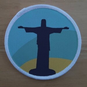Puede incluir: Un parche circular blanco con un fondo azul y amarillo que presenta una silueta negra de la estatua del Cristo Redentor en Río de Janeiro, Brasil.