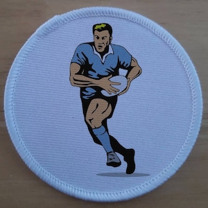 Puede incluir: Un parche redondo blanco con una ilustración azul y blanca de un jugador de rugby corriendo con el balón. El jugador lleva una camiseta azul y pantalones cortos negros.
