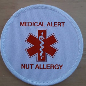 Può includere: Cerotto di allerta medica rotondo bianco con testo rosso e un simbolo rosso di stella della vita. Il testo recita "MEDICAL ALERT" e "NUT ALLERGY".