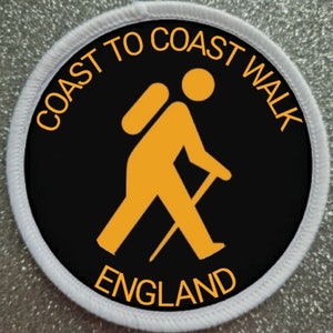 May include: A black and white round patch with a white border. The patch has a yellow silhouette of a person walking with a backpack and a walking stick. The text "COAST TO COAST WALK ENGLAND" is written in yellow around the silhouette.