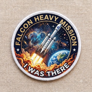 Op de afbeelding: Een ronde geborduurde patch met een Falcon Heavy raket die de ruimte in gaat, met een nevelachtergrond en de aarde zichtbaar. De tekst FALCON HEAVY MISSION en I WAS THERE staat in dikke gele en witte letters met een witte rand.