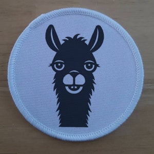 May include: A white round fabric patch with a black cartoon llama face. The llama has a happy expression with its mouth open and teeth showing.