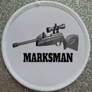 Puede incluir: Parche ovalado blanco con un rifle de aire comprimido negro y plateado con una mira telescópica y la palabra "MARKSMAN" en letras negras.