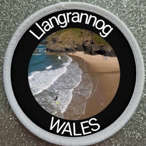 May include: A white round patch with a black circle in the center. The black circle has a white border and the text "Llangrannog WALES" in white. The center of the circle features a photo of a beach with waves crashing on the shore.