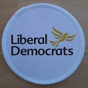 Puede incluir: Un parche redondo de tela blanca con un logotipo de pájaro amarillo y el texto "Liberal Democrats" en negro.