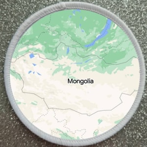 May include: A white round patch with a green and white map of Mongolia. The map shows the country's borders and major geographical features. The word "Mongolia" is printed in black text in the center of the patch.