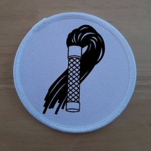 May include: A white round patch with a black and white illustration of a whip. The whip is coiled and has a handle with a grid pattern.