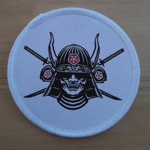 Op de afbeelding: Een ronde witte patch met een zwart-witte illustratie van een samoerai-krijger die een helm draagt met rode bloemen. Twee zwaarden zijn gekruist achter het hoofd van de krijger.