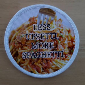 Puede incluir: Un parche redondo blanco con un tenedor y un plato de espaguetis con salsa de carne. El texto en el parche dice "Menos Upsetti, Más Spaghetti."