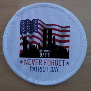 以下が含まれることがあります： 赤、白、青のアメリカ国旗が風になびいている白い丸いパッチ。旗は、ツインタワーを含むニューヨーク市のスカイラインの黒いシルエットの前にあります。「SEPTEMBER 9/11 NEVER FORGET PATRIOT DAY」というテキストが画像の下に印刷されています。