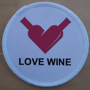 Puede incluir: Parche redondo blanco con un corazón rosa formado por dos botellas de vino. El texto "LOVE WINE" está impreso debajo del corazón.