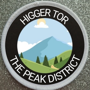 Op de afbeelding: Een witte en zwarte ronde patch met de tekst "Higger Tor The Peak District" en een cartoonafbeelding van een berg met een blauwe lucht en een witte wolk.