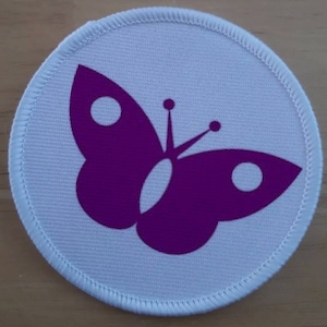 May include: A white round fabric patch with a purple butterfly design. The butterfly has two large wings and a small body with two small circles on each wing.