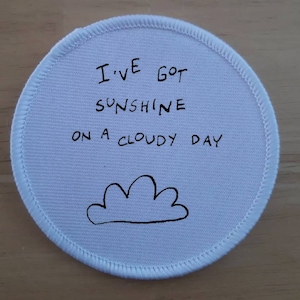 May include: White round fabric patch with black stitching around the edge. The patch has black handwritten text that reads "I've got sunshine on a cloudy day" and a simple black drawing of a cloud.