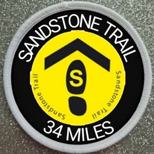 May include: A white and black circular patch with the text "Sandstone Trail" in white lettering around a yellow circle. The yellow circle has a black silhouette of a footprint with the letter "S" inside. The text "34 Miles" is printed in white below the yellow circle.