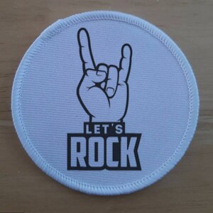 Pode incluir: Patch redondo de tecido branco com uma ilustração em preto e branco de uma mão fazendo o gesto de rock and roll. O texto "Let's Rock" está impresso em preto abaixo da mão.