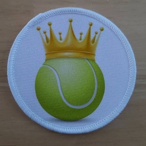 May include: A white round patch with a green tennis ball wearing a gold crown.